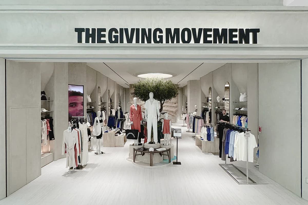 Dominic Nowell-Barnes, founder of The Giving Movement, photographed in front of a mood board displaying brand imagery and the words 'minimalism' and 'futurism', with a TGM logo and a model wearing the brand's signature olive tracksuit.
