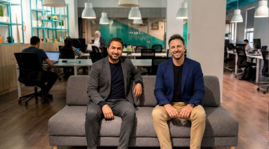 Aumet co-founders Yahya Aqel and Adel Haddad standing inside a modern pharmacy or hospital supply room, with a laptop showing the Aumet procurement dashboard.