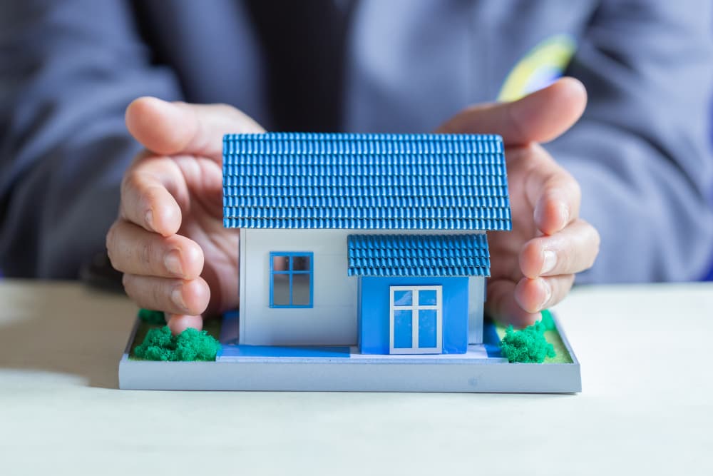 Hands cupped protectively around a small model house with a blue tiled roof on a tabletop, symbolizing home ownership and housing loan protection in Lebanon