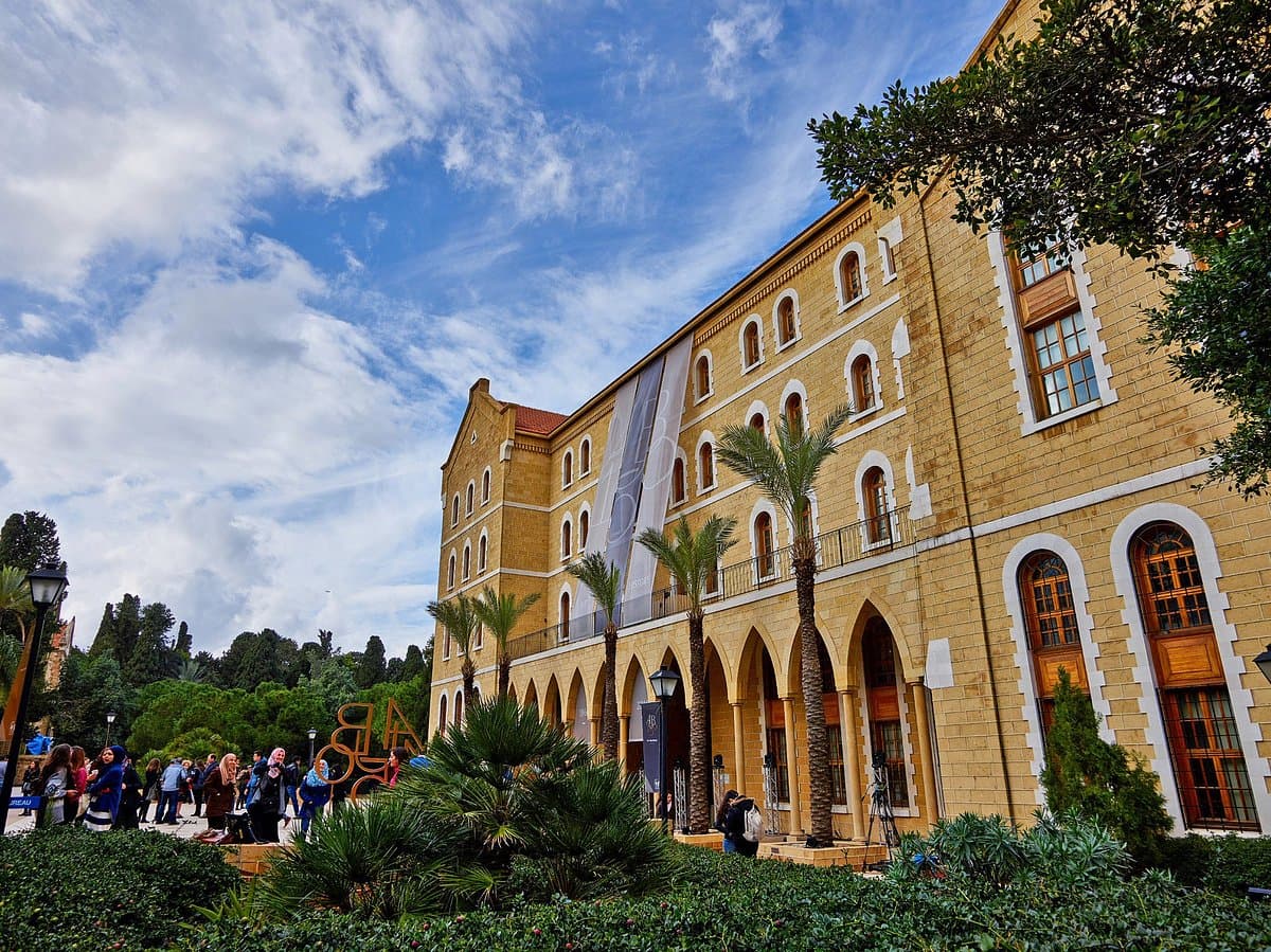 A sunny view of the historic yellow sandstone College Hall building on the American University of Beirut campus, with arched walkways, tall palm trees, green gardens, and students gathered near the entrance under a blue sky.