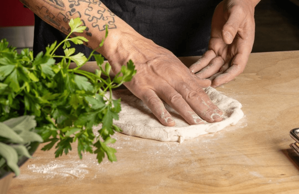 A chef in a black apron with a tattooed forearm shapes pizza dough by hand on a floured wooden surface, with fresh parsley and sage in the foreground.
