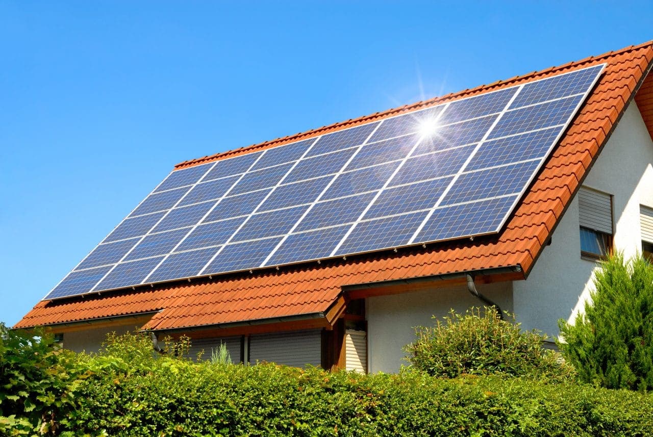Solar photovoltaic panels mounted on the red tiled roof of a modern house, with the sun flaring off the panels against a clear blue sky and green hedges in the foreground.