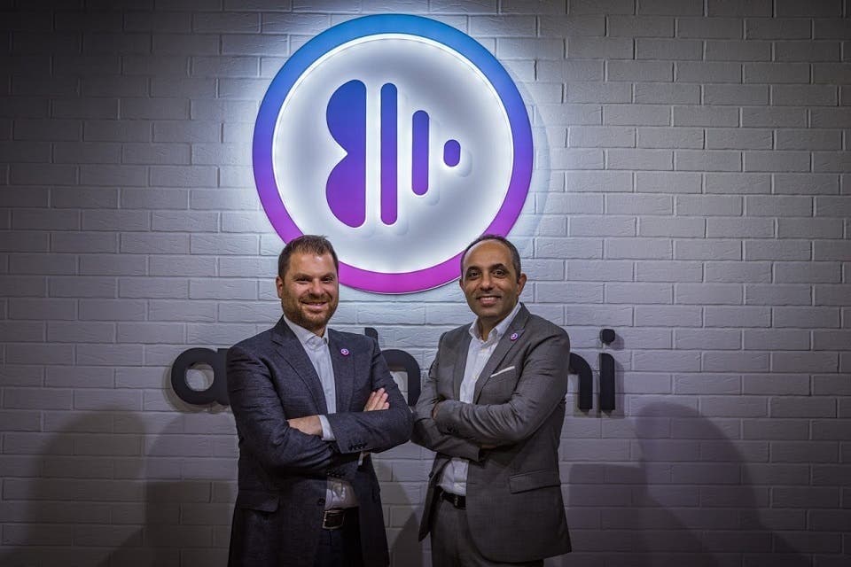 Anghami co-founders Eddy Maroun and Elie Habib standing with arms crossed in front of the glowing Anghami logo on a white brick wall