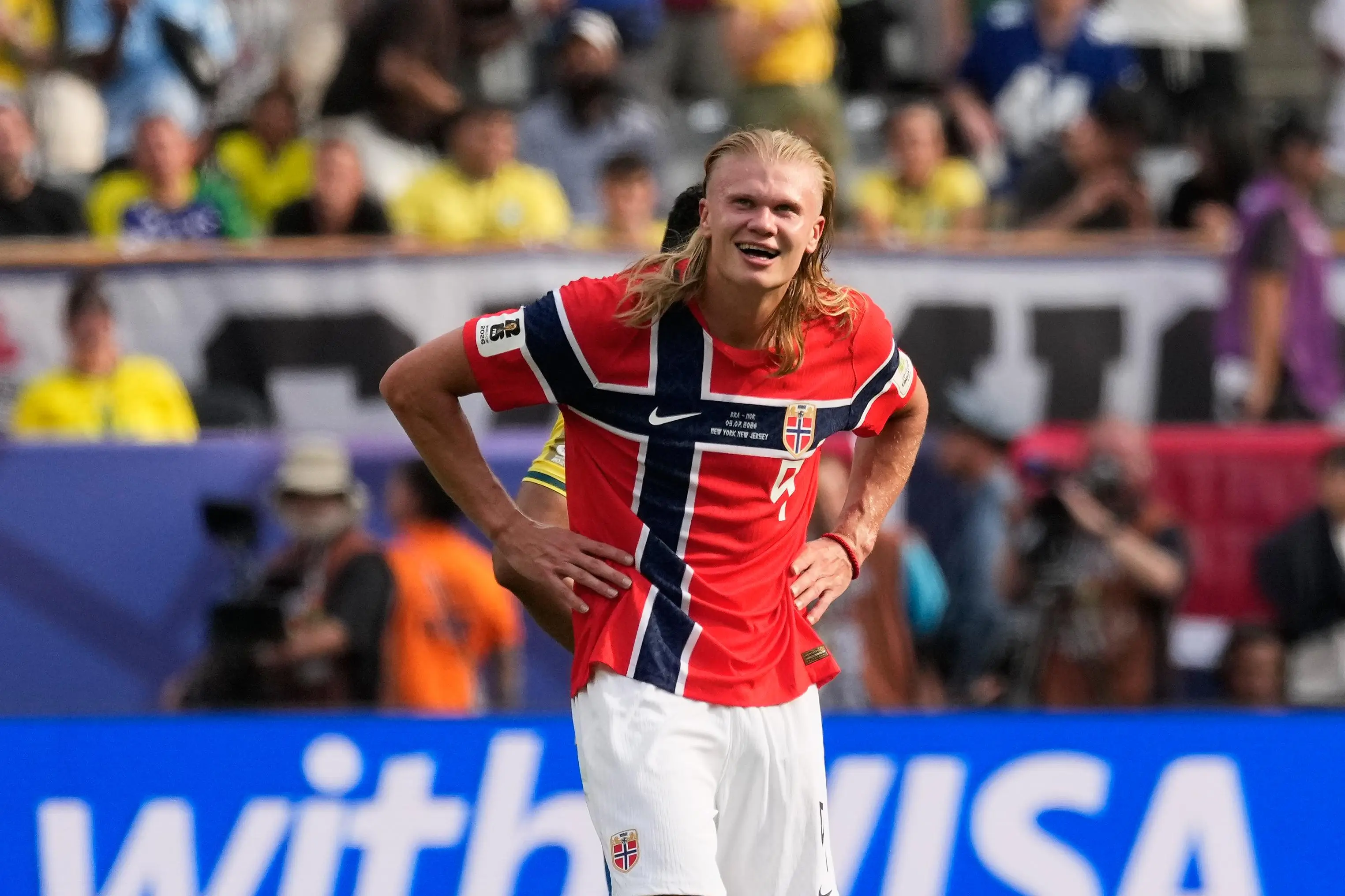 Erling Haaland in Norway's red home kit with the blue-and-white flag cross, hands on hips and smiling on the pitch during the Brazil vs Norway 2026 World Cup match in New York, New Jersey, with a crowd blurred behind him.