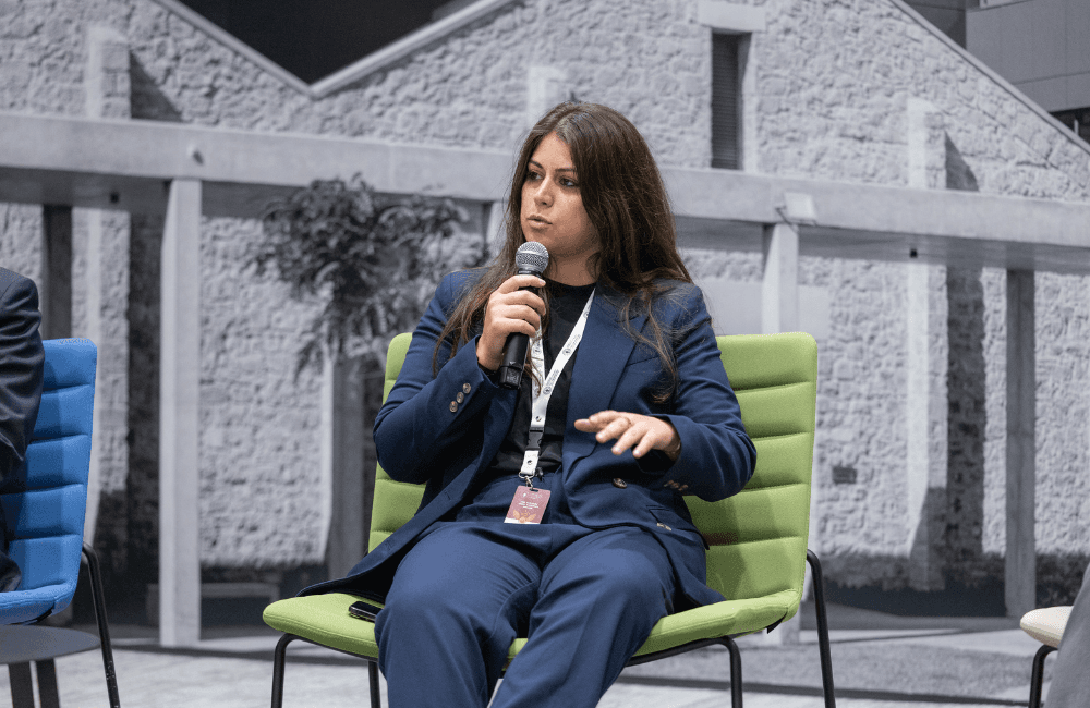 Sandra Helou sits in a green chair wearing a navy blue suit, holding a microphone as she speaks on a panel, in front of a large black-and-white photo of a stone building.