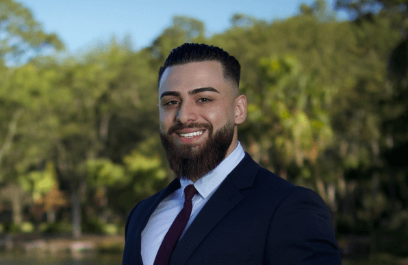 Kevin Kallassy, a young real estate agent with a dark beard, smiling in a navy suit, white shirt, and dark red tie, photographed outdoors in front of green trees.