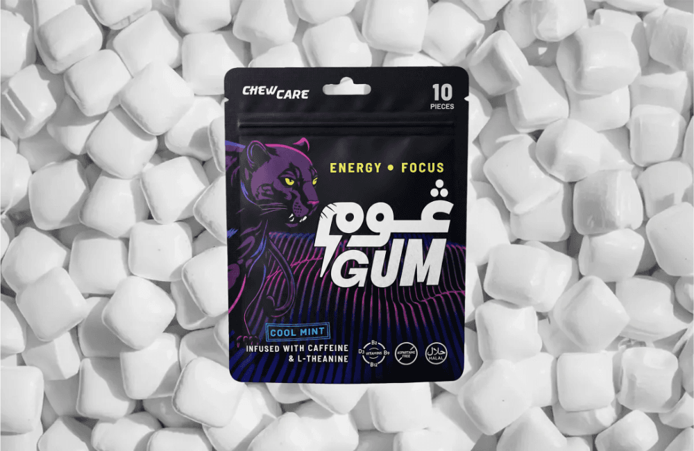 Black Goom energy gum pouch by ChewCare, labelled "10 pieces" and "Cool Mint," with a purple and pink neon panther graphic and the Arabic word غوم beside "GUM." The front reads "Energy, Focus" and "Infused with caffeine and L-theanine," with B2, B6, B12 and D3 vitamin badges plus aspartame-free and halal marks. The pouch rests on a bed of white gum pieces.