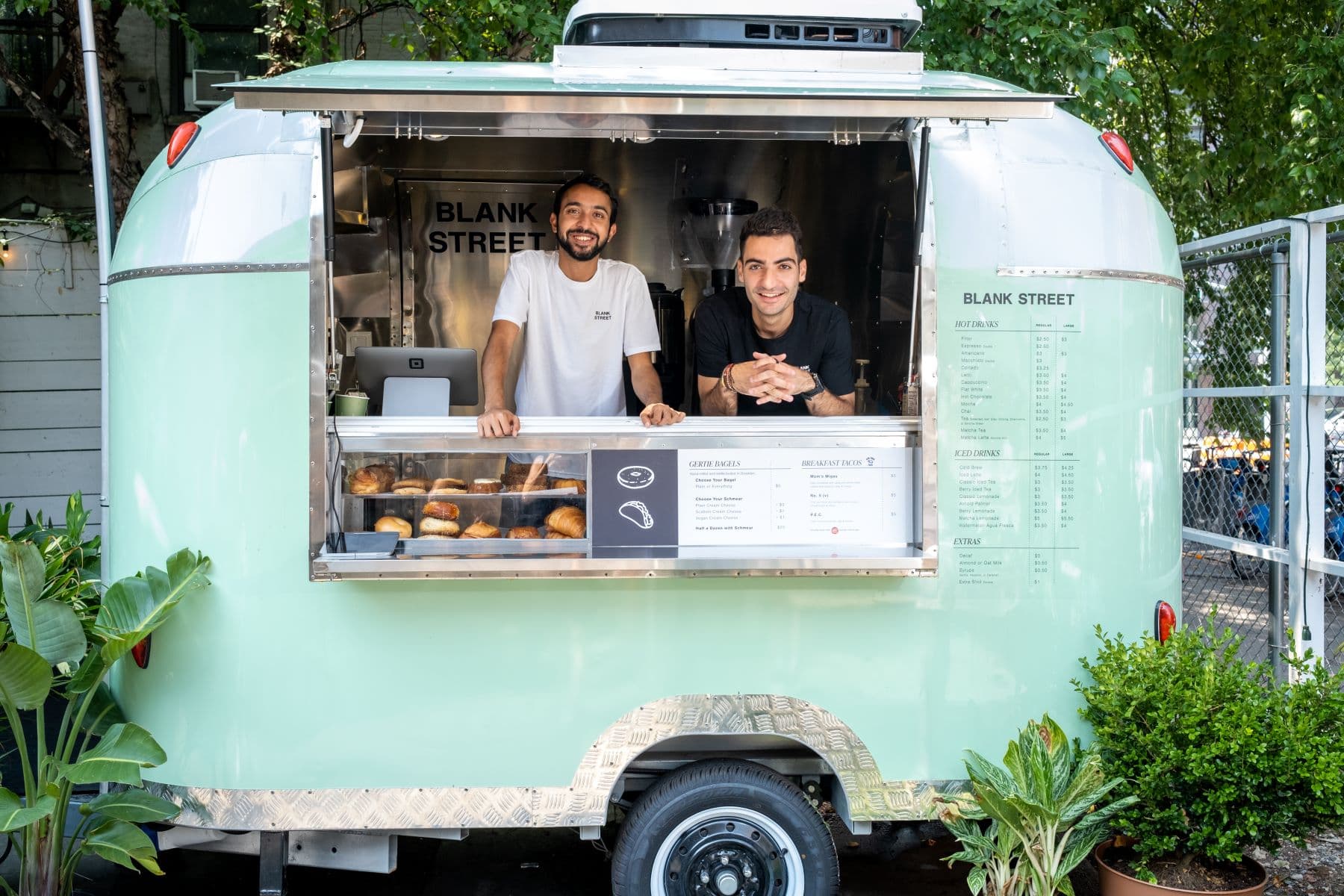 Two Blank Street co-founders standing at the service window of the brand's mint green trailer coffee cart in Brooklyn, with a pastry case and menu board in front of them.