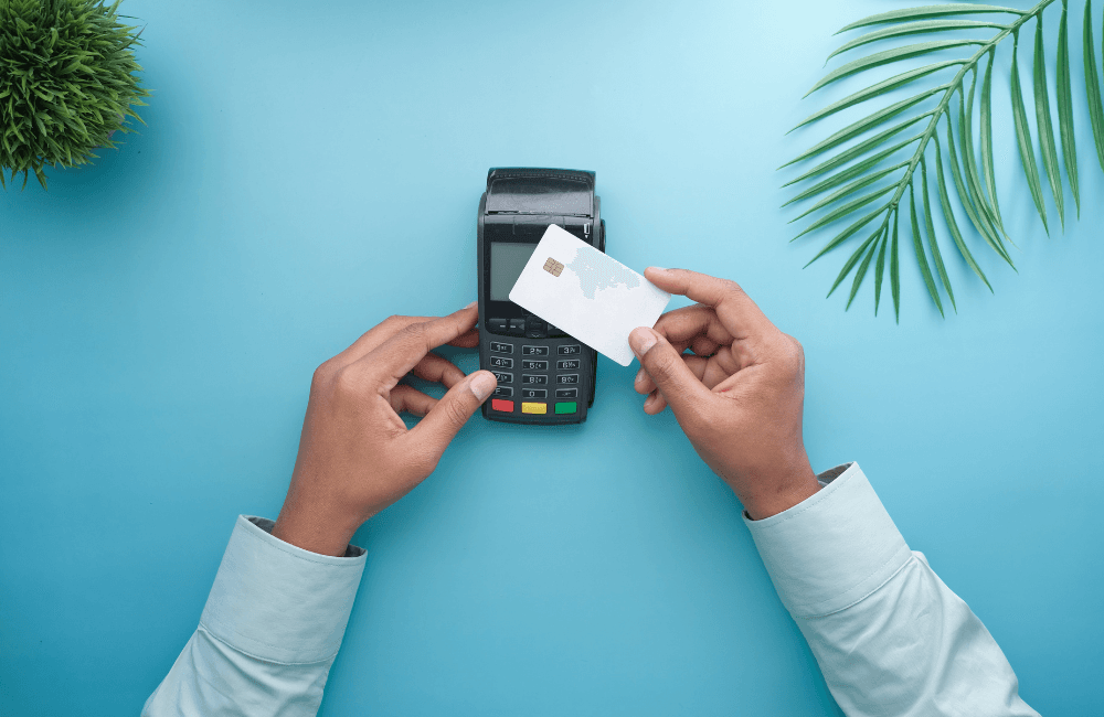 Overhead view of hands holding a blank white chip card against a black point of sale terminal on a light blue background, framed by a small plant and a palm leaf.