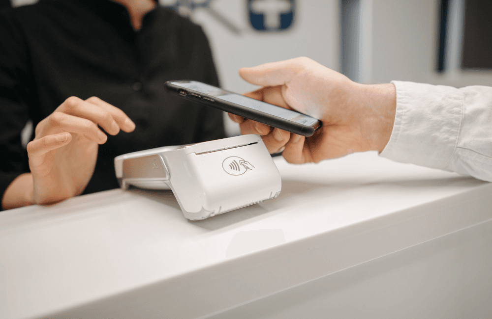 A customer holds a smartphone over a white contactless card reader on a shop counter while the cashier waits to complete the payment.