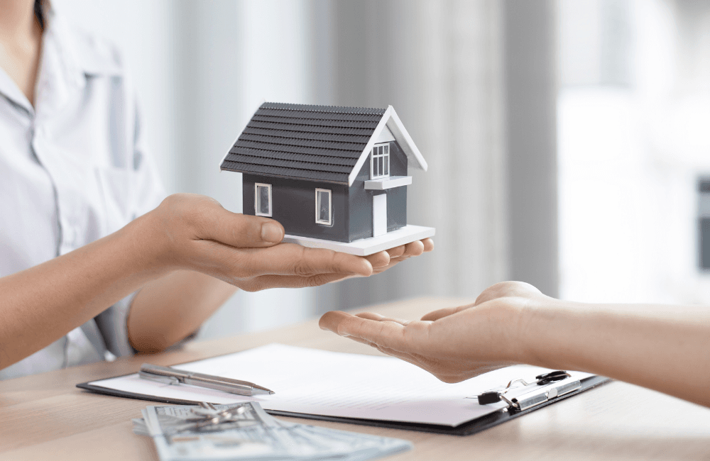A person hands a small grey model house to another person's open palm across a desk with a clipboard, a pen, house keys and US dollar bills.
