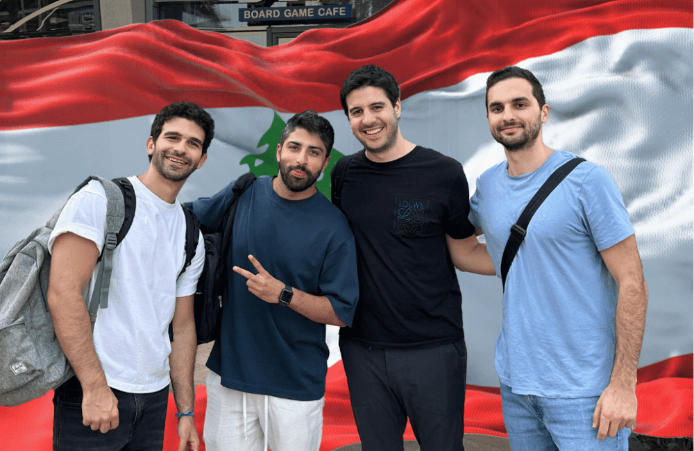 The four Panic Zones co-founders smiling together in front of a large Lebanese flag outside a board game cafe.