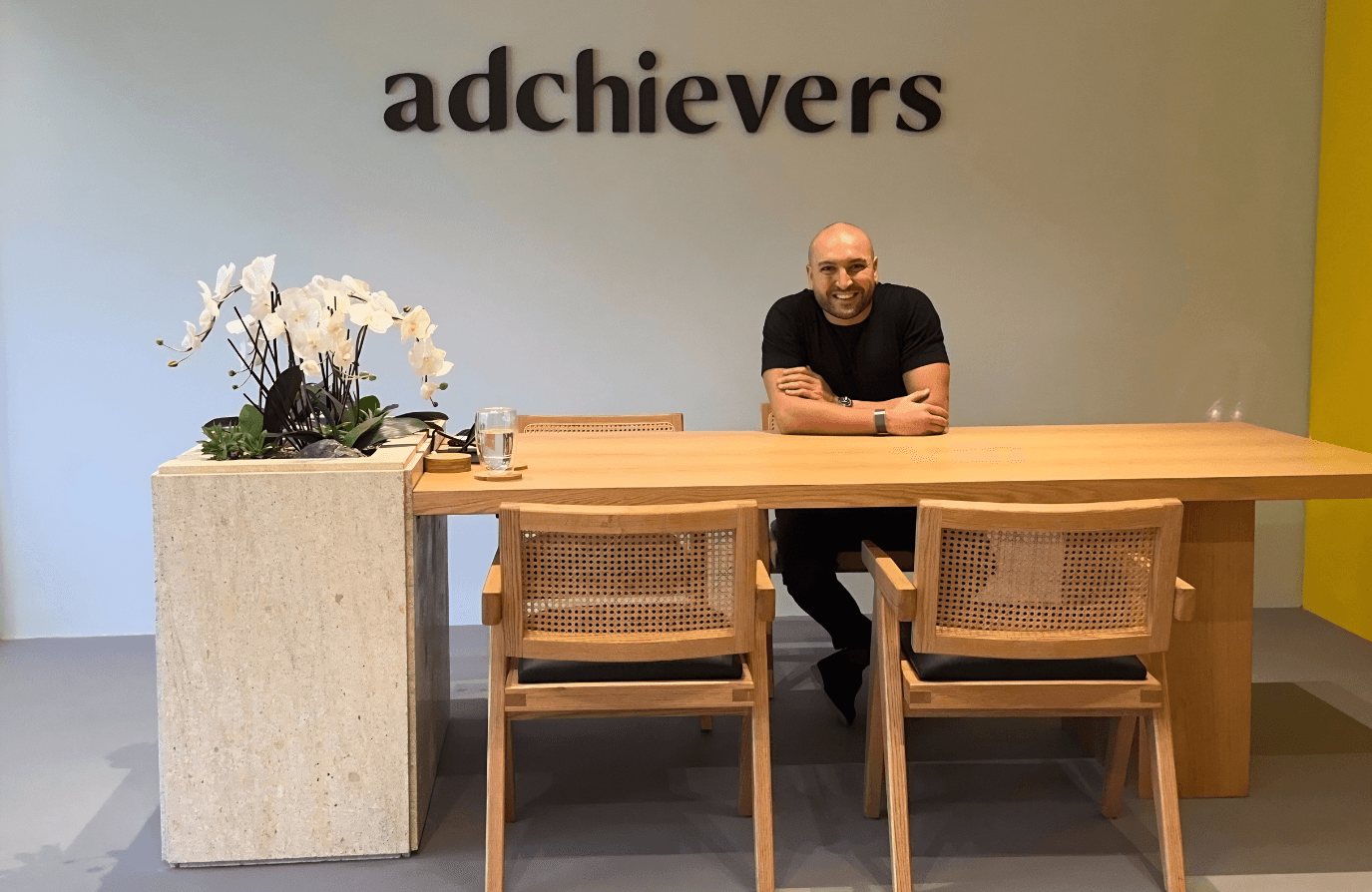 Elias Saliba, founder of Adchievers, sitting at a wooden meeting table under the Adchievers logo in the agency's office