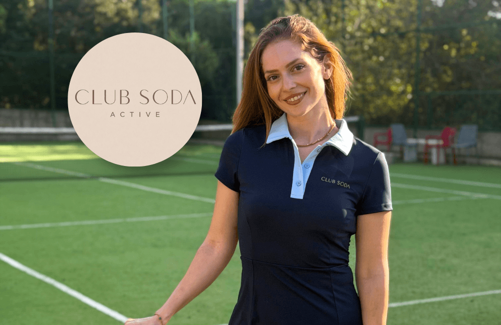 A woman smiles on a green tennis court, wearing a navy CLUB SODΛ ACTIVE tennis dress with a light blue collar. Beside her is a circular beige CLUB SODΛ ACTIVE logo.