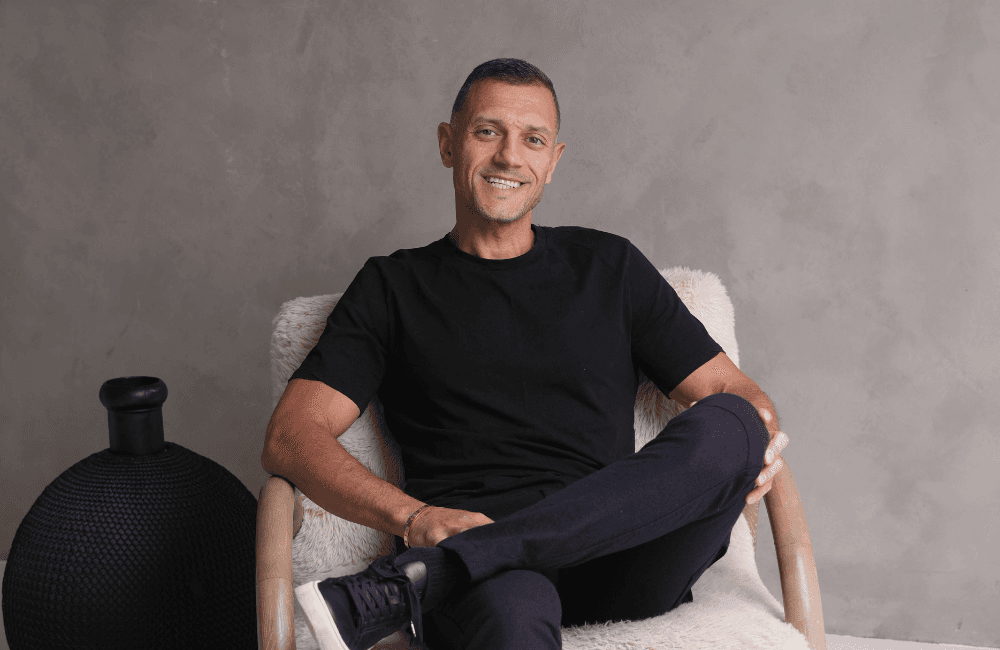 Sam Kamra, founder of Listing.ca, smiling while seated in a white sheepskin armchair, wearing a black T-shirt and dark trousers, against a grey textured wall