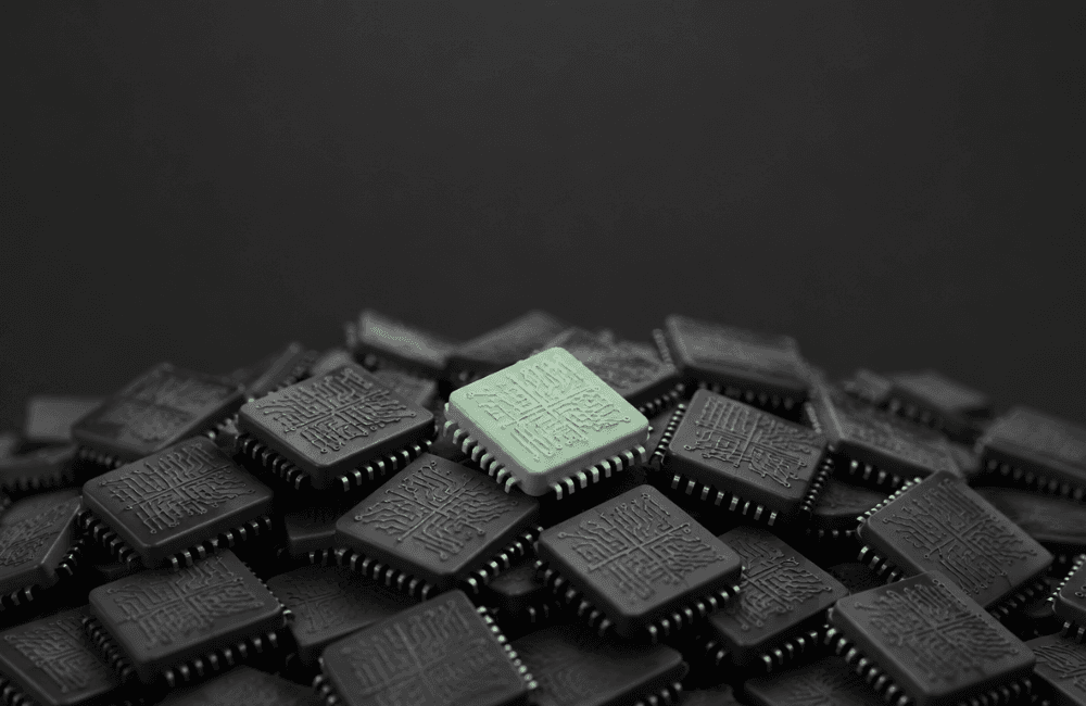 A single mint green microchip sits on top of a large pile of dark grey microchips against a plain black background.