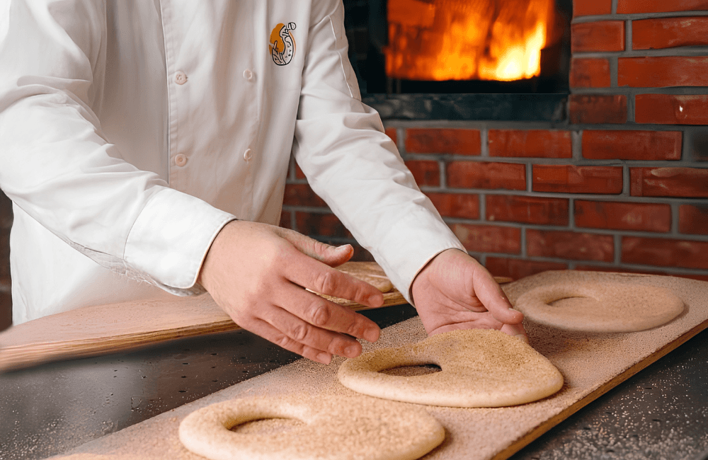 A Kaaki W Nos baker in a white chef’s jacket shapes raw sesame kaak dough on a wooden board in front of a brick oven with an open fire.
